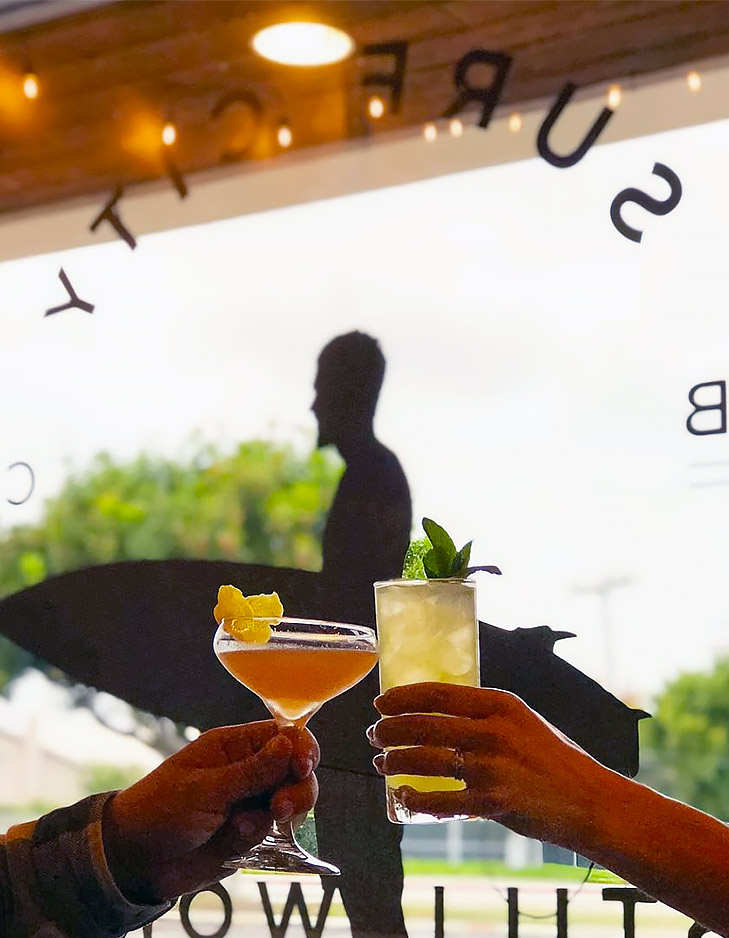 image of two hands toasting with cocktails in front of surf city still works window